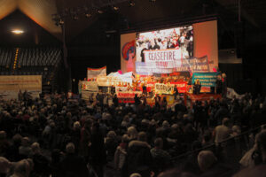 Foto der Saalkundgebung "Manifestation für einen gerechten Frieden in Nahost" der Rosa-Luxemburg-Konferenz 2024 in Berlin. Auffällig ist die Bühne voller Genossinnen und Genossen, die Transparente und Banner hochhalten gegen Krieg und gegen Nato. Ein Banner sagt "Palästina sagt: ich war, ich in, ich werde sein" in Anspielung auf Israels Völkermord im Gazastreifen.