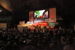 Foto der Saalkundgebung "Manifestation für einen gerechten Frieden in Nahost" der Rosa-Luxemburg-Konferenz 2024 in Berlin. Auffällig ist die Bühne voller Genossinnen und Genossen, die Transparente und Banner hochhalten gegen Krieg und gegen Nato. Ein Banner sagt "Palästina sagt: ich war, ich in, ich werde sein" in Anspielung auf Israels Völkermord im Gazastreifen.